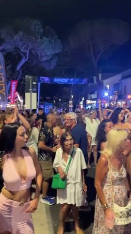 The video shows a crowded street at night, likely during a festival or event, with many people dancing and enjoying themselves. The street is lined with buildings, some adorned with lights. A banner with the text "WRS" is visible across the street. People of various ages are present, dressed in casual summer attire. Some individuals are holding up their hands or phones, capturing the moment. The atmosphere appears festive and lively, with a sense of community celebration.