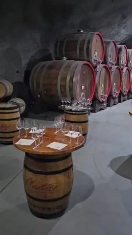 The video pans across a dimly lit wine cellar. Rows of large wooden barrels, some stacked two high, line the arched stone walls. In the foreground, several wine barrels have been repurposed as small tables, each topped with a round wooden surface and set with multiple wine glasses. Some of the barrels have visible branding, including "Plantaze" on the barrel in the lower left. The floor is concrete and appears clean. The lighting is subdued, with spotlights illuminating some of the barrels. No people are visible in the video.