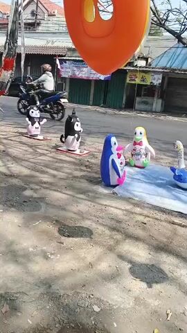 The video captures an outdoor scene. In the foreground, a variety of inflatable toys are displayed on a white sheet laid on the ground. These include several penguin-shaped inflatables of different colors (blue, red, green, purple, and yellow with pink accents) and sizes, as well as a blue inflatable resembling a whale with its tail raised. Behind the toys, a street is visible with light traffic. Several motorcycles are parked on the side of the road, and at least two are in motion, with riders. A man in a black jacket and helmet rides a motorcycle from left to right across the frame. Further in the background, there are buildings, one with a prominent red sign that reads "erafone." Trees and the sky are also visible. The lighting suggests it is daytime.