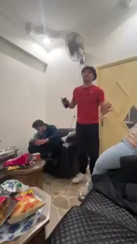 Three people are in a room. One man stands in the center of the room wearing a red t-shirt, black pants, and white sneakers. He gestures with his arms, appearing to dance. A second man sits on a black chair, looking down at an object in his hands. He wears a blue and black jacket. A third person sits on a black couch, partially visible, wearing light blue jeans and a dark jacket. On a small, carved wooden table in front of the couch are various snacks. A television sits on a wooden stand in the background. The room has tiled floors and white walls. There is a ceiling fan, a door in the background, and a small space heater. The man in the red shirt walks to the left and dances behind a man wearing a white hoodie and black pants.