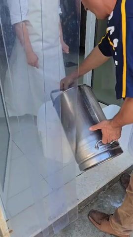 From a partially obscured perspective behind a tall, clear plastic panel, a person wearing a white apron stands next to a white bucket lined with a white cloth. Another person, wearing a black shirt with yellow stripes on the sleeves, pours liquid from a large stainless-steel bucket into the white bucket. The floor is covered with white, square tiles. Another white bucket is visible in the background to the right. The lighting appears to be bright, possibly indoors.