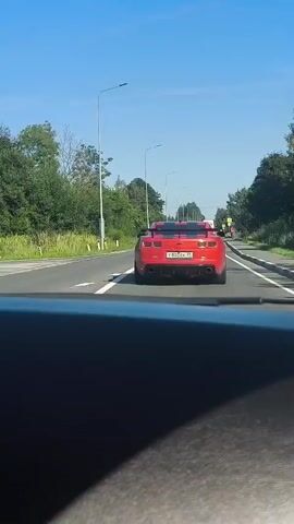 This video captures a view from inside a vehicle driving on a paved road under a clear, bright blue sky. The time of day appears to be midday. The road is flanked by lush green trees and foliage on both sides, suggesting a rural or suburban setting. Sunlight creates distinct shadows, indicating sunny weather.

The primary focus is a bright red Chevrolet Camaro convertible driving ahead in the same lane. The car has black racing stripes on its hood and trunk, and its taillights are illuminated. The license plate is visible and reads "T800EK 39".

Other vehicles are present on the road: a dark grey car is visible in the left lane in an earlier frame, and a white van is seen in the distance ahead. In the right lane, further ahead, a person in a red shirt is walking along the side of the road. In the background, there are lampposts and traffic signs.

The scene is one of typical road travel, possibly on a highway or a well-maintained country road, with the distinct visual of the red Camaro as the main subject of interest. The overall mood is calm and observational, with no indication of urgency or distress. The location context of Откосово, Russia, points to the geographical setting of this drive.