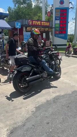 A person sits on a motorcycle in front of a gas station. The person is wearing a helmet with rainbow stripes and a black jacket. The gas station has a sign that says "FIVE STAR". There are other motorcycles parked around. The gas prices are displayed on a digital screen.