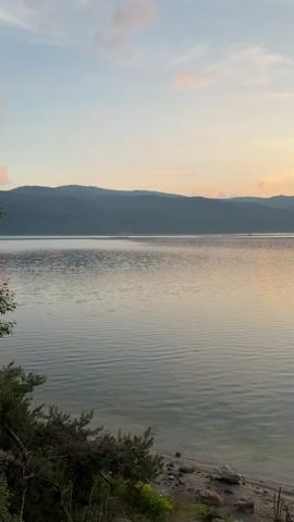 The video shows a time-lapse of a sunset over a wide lake or body of water, framed by a dark tree branch at the top right. The sky transitions from a soft blue with wispy clouds to vibrant hues of orange and yellow as the sun dips below a distant mountain range on the right. The calm water reflects these colors, with gentle ripples visible across its surface. The foreground features a rocky shore with some sparse green and yellow vegetation. Dark, silhouetted mountains stretch across the horizon in the background. As the video progresses, the sun becomes more prominent on the right, illuminating the clouds and water with a warm glow.