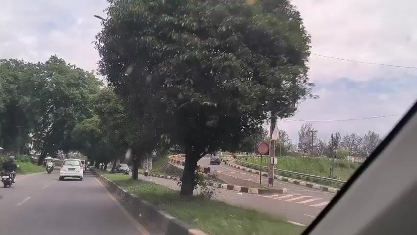 The video shows a view from inside a car driving on a road. The road is lined with trees on the left side and a grassy area with a lower road on the right. There are cars and motorcycles moving in both directions on the road. The sky is overcast.