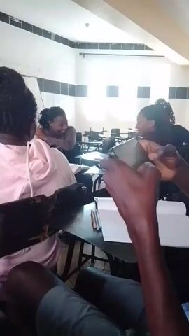 The video shows a classroom setting with multiple students seated at desks. The room is brightly lit with natural light coming through several windows. The walls are painted in a light color, with some sections highlighted in darker tones. Most students are Black.  They are either conversing with each other or focusing on their phones. Some have notebooks and pens on their desks. The camera appears to be held by someone seated among the students, offering a first-person perspective. The camera moves throughout the classroom.