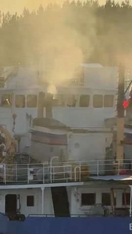 The video shows a large ship sailing on a body of water, possibly a river or canal. Smoke is emanating from one of the ship's smokestacks. The ship has a multi-story white superstructure with multiple windows. There are flags on the ship, including what appears to be a Russian flag. The ship's hull is primarily blue. The background consists of a line of trees and a sky with clouds, illuminated by what appears to be sunset or sunrise light. The overall impression is of a working vessel in transit.