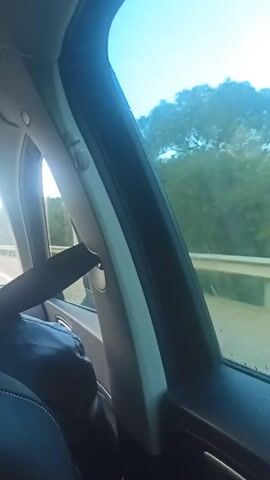 The view from a car window shows trees passing by. The sky is blue with a few clouds. The camera is focused on the window frame, which is white with black trim. A seatbelt is visible in the lower left corner. The car is traveling on a road with a guardrail.
