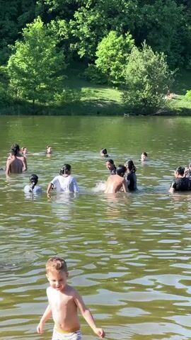 A group of approximately fifteen people are standing and wading in a lake, including a small child in the foreground facing the camera. The child, wearing swimming trunks, is smiling and waves with their right hand.  Some people in the lake are dressed in clothing, while others are shirtless or in bathing suits.  They are interacting with each other, splashing and talking.  The lake is surrounded by green trees and grass, with sunlight dappling the water's surface.  One person is visible on the grassy bank behind the trees.