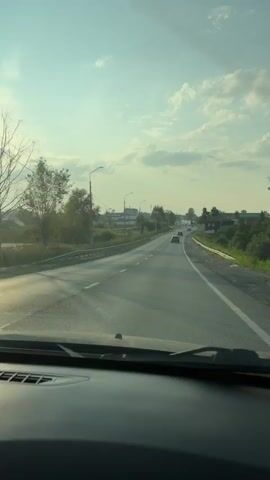The video shows a first-person perspective from inside a vehicle traveling on a paved road. The road has lane markings, with a white line on the right and a broken white line on the left. There are streetlights and power lines visible along the side of the road. On the left, there is a metal guardrail and some trees and vegetation. On the right, there are more trees and some buildings in the distance. Several other vehicles, including cars and at least one motorcycle, are also traveling on the road in the same direction. The sky is light blue with some scattered clouds, and the sun appears to be low in the sky, casting long shadows. The overall environment suggests a rural or suburban setting during daylight hours.