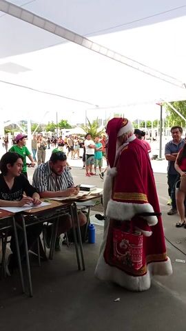 This video, filmed in Ñuñoa, Chile, captures an outdoor festive scene. In the foreground, Santa Claus is prominently featured, dressed in a traditional red and white suit with gold trim, a black belt, and a long white beard. He is holding a red bag adorned with the words "MERRY CHRISTMAS" and an image of Santa, resting it on a table. Santa appears to be leaning forward, perhaps interacting with the people seated at the table.

Two individuals are seated at a wooden table in front of Santa. The person closer to the camera wears a black t-shirt and has short dark hair, focusing on papers in front of them, holding a pen. Next to them, another person in a light blue and white striped shirt is also engaged with papers on the table, holding a pen and looking towards Santa. A blue water bottle is visible under the table.

The setting is outdoors, beneath a large white canopy or tent structure, which provides shade. The ground appears to be paved concrete. In the background, numerous people are visible, some standing in lines or groups, suggesting a public event or gathering. Trees and parts of buildings can be seen beyond the crowd, indicating an urban or park-like environment. The lighting suggests it is daytime. The overall atmosphere appears calm and festive, with Santa as the central figure of interaction.