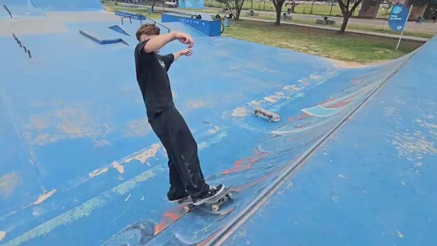 The video shows three people skateboarding at a skate park. The skate park has a blue surface with graffiti. The first person shown is wearing a black shirt and black pants and slides down a ramp on their skateboard. The second person is wearing a black shirt and black pants and is skateboarding around the park. The third person is wearing a black shirt, black shorts, white socks, and a baseball cap. He tries to ride a ramp on his skateboard but the skateboard slips out from under him.