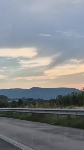The media file depicts a scenic view from a moving vehicle, likely on a highway, as indicated by the asphalt road and guardrail in the foreground. The scene is set in a rural or mountainous region, with rolling hills and distant mountains forming the backdrop. The time of day is either sunrise or sunset, suggested by the warm hues of orange and pink present in the sky, contrasting with the grey clouds. The weather appears partly cloudy, with the sun peeking through a break in the clouds, creating a bright, luminous effect. The overall atmosphere is serene and tranquil. There are no people or identifiable objects other than the natural landscape and the roadside infrastructure. No visible text is present in the media file. The location is identified as Peshterna, Bulgaria.