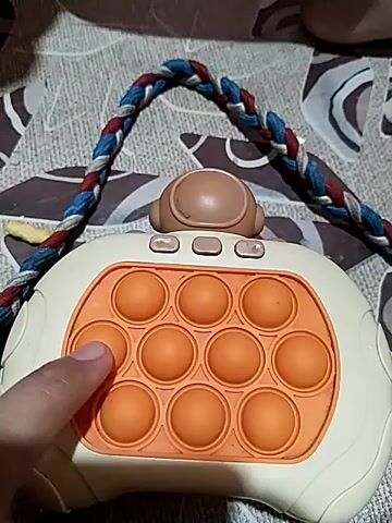 A close-up video shows a hand interacting with a pop-it toy. The toy is light-colored with an orange pop-it section containing nine circular bubbles arranged in a 3x3 grid. A braided strap made of blue, red, and white cords is attached to the top of the toy. The background is a patterned textile, possibly a tablecloth. The hand, with visible fingernails and skin texture, presses down on the pop-it bubbles, making them click and indent. The video focuses on the tactile experience of popping the bubbles.