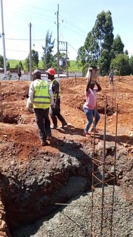 The video shows construction workers pouring concrete into a foundation. Three people are visible near the foundation hole. Two of them are wearing reflective vests and carrying buckets, while the third is carrying a bucket on their head. The foundation hole is filled with wet concrete. Steel rebar is arranged in a grid pattern at the bottom of the hole and extending upward, forming a cage structure. The surrounding ground is red dirt. Power lines and green trees are visible in the background under a bright blue sky. The workers are actively pouring more concrete into the foundation.