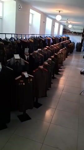 The video pans across a room filled with rows of uniforms displayed on racks. The uniforms appear to be military or ceremonial in nature, with many featuring distinctive collars, epaulets, and medals. Some are on mannequins, while others are hanging directly from the racks. In the background, shelves line the walls, filled with what appear to be hats or helmets. The room is well-lit, with ceiling lights visible, and the floor is tiled. The overall impression is that of a museum or a collection of historical uniforms.