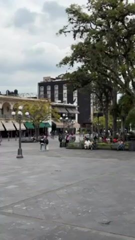 The video shows a public square on an overcast day. People are walking around or sitting on benches in the park. There are buildings visible in the background, as well as trees and lampposts. A person in a red shirt and white shorts walks across the square in the foreground. The ground is paved with stone tiles.