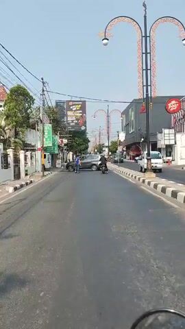 The video shows a street scene with a gray sedan turning left in front of a motorcycle. A person in a blue shirt stands on the sidewalk near the turning car. A Pizza Hut restaurant is visible on the right side of the street. The sky is clear and the weather appears to be sunny. The camera appears to be mounted on a motorcycle, indicating it is a first-person perspective.