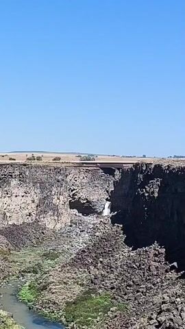 A static, high-angle shot captures a wide canyon with a river at the bottom. Towering cliffs, predominantly composed of dark, jagged rocks, flank the canyon. A modest waterfall cascades down the cliff face in the distance. Above the canyon, a bridge spans the gap, with a few scattered trees and a vast, flat expanse of dry land visible on the horizon under a clear blue sky. The foreground is dominated by a dense collection of dark, boulder-like rocks.