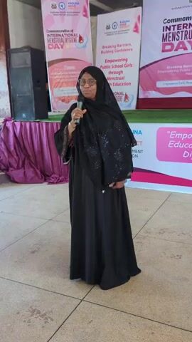 A woman in a black hijab and dress stands at a podium, holding a microphone and speaking. She wears glasses and appears to be addressing an audience. Behind her, several banners are displayed, indicating a public event. The banners feature text in English, with the Kaduna State Government and Ministry of Education being prominent. One banner reads "Commemoration of INTERNATIONAL MENSTRUAL HYGIENE DAY" and lists the theme as "Breaking Barriers, Building Confidence: Empowering Public School Girls through Menstrual Hygiene Education." Another banner states "KADUNA STATE GOVERNMENT MINISTRY OF EDUCATION" and "Empowering Public School Girls through Menstrual Hygiene Education."

The setting appears to be outdoors or in a semi-covered area with a tiled floor. To the left of the speaker, a large black speaker is visible on a platform draped with a purple cloth. Steps are also visible to the far left. The lighting suggests daytime, possibly morning or afternoon. The woman's posture and expression are neutral, indicating a formal presentation. The overall scene depicts an awareness or educational event related to menstrual hygiene in Nigeria.