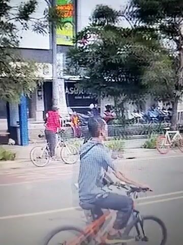 The video shows a street scene with several people. In the foreground, a person rides a bicycle on the right side of the road. The road has two lanes marked by white lines. On the left side of the road, there is a sidewalk with trees and buildings. Some people are walking on the sidewalk, and others are standing near the road. In the background, there are more trees and buildings, as well as a bridge or overpass with banners on it. Some people are seen holding up phones, possibly recording or taking pictures. The overall atmosphere appears to be that of a public gathering or event. The quality of the video is somewhat blurry and pixelated, making it difficult to discern fine details.