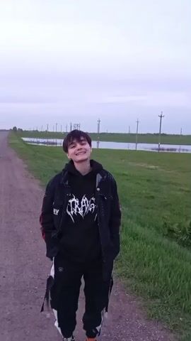 A person with short, dark hair stands on a dirt road. They are wearing a black hoodie with a white graphic, a black jacket with red accents, and black pants with white straps. The person is smiling and looking at the camera. The background shows a flat, grassy landscape with standing water and telephone poles in the distance. The sky is overcast. The camera appears to be handheld and moves slightly.