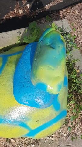 The video showcases a cluster of painted rocks, arranged to resemble a whimsical creature, possibly a turtle or an insect, resting outdoors. The rocks are vividly colored with alternating patterns of bright yellow and blue spray paint. The largest rock, painted yellow with blue swirling lines, forms the base and likely represents the body. Two smaller rocks, one primarily blue and the other a mix of blue and green, are positioned to suggest a head and possibly a limb. The scene is set in a garden or natural outdoor setting, with soil, dry leaves, and green vegetation visible around the rocks. The lighting suggests it is daytime, with sunlight casting highlights on the painted surfaces, indicating a sunny or bright day. The overall impression is one of playful, homemade garden art. There is no visible text in the video.