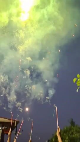 A group of people are holding sparklers in the air as fireworks explode in the night sky. The people are smiling and appear to be enjoying themselves. The fireworks are red and white and are creating a lot of smoke. The sky is dark and there are some trees in the background.