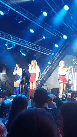The video shows a live performance by three female singers on a stage with bright blue and purple lights. They are all wearing matching outfits consisting of white tops and red skirts, with the singer on the left also wearing blue boots. The audience is visible in the foreground, with several people holding up phones to record the performance. Behind the singers, a drummer can be seen playing drums. The stage also has a large screen with glowing text that appears to read "DADADAK". The overall atmosphere appears to be energetic and exciting.