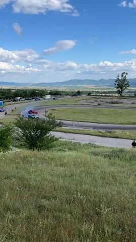 At 00:00, two cars are visible on a race track, one red and one blue and white. The red car is on the left, drifting and producing white smoke. The blue and white car is on the right, also drifting. The track is surrounded by green grass and is in a rural area with mountains in the background. The sky is blue with scattered clouds. At 00:01, the blue and white car has moved further to the right, still drifting. At 00:02, the red car is still drifting on the left, and the blue and white car is drifting on the right. Two people are standing in the grass in the foreground, watching the cars. At 00:03, the red car continues to drift, producing a large amount of smoke. The blue and white car is now further ahead and to the right, still drifting. At 00:04, the two cars are further along the track, with the red car now behind the blue and white car. Both are still drifting. At 00:05, the cars continue to drift around a bend in the track.