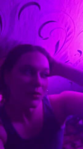 A young woman with brown hair and makeup, including blue eyeshadow and red lipstick, is shown in a dimly lit room with purple lighting. She is wearing a grey tank top and large hoop earrings. The background features textured wallpaper with a swirling pattern. The woman is looking off-camera and touching her hair, then she turns to face the camera and speaks, gesturing with her hand.