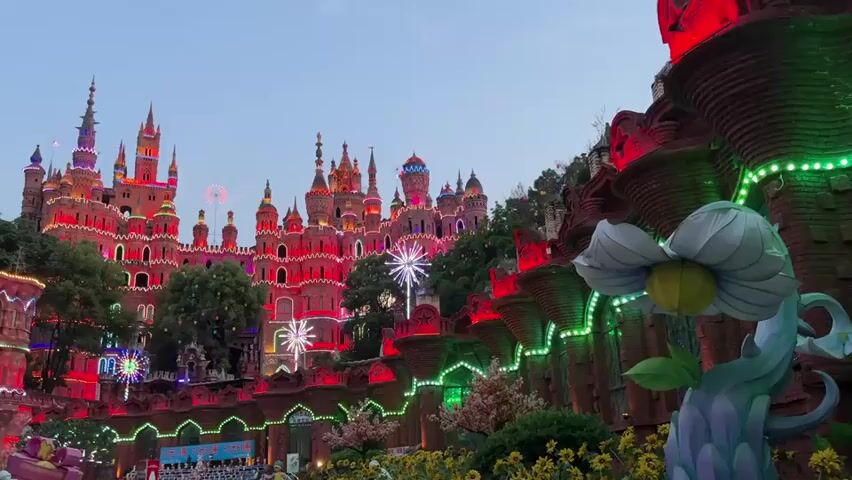 The video shows a large, colorful castle illuminated with various colored lights, including red, green, blue, and white. The castle has many towers and spires. There are trees surrounding the castle. In the foreground, there's a large, stylized white flower sculpture, also lit up. There appears to be a gathering of people in front of the castle. The sky is a pale blue. The overall scene is vibrant and festive.