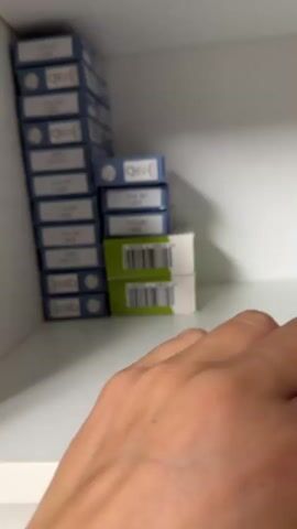 A hand reaches into a white shelf and picks up a blue box of anchovies. The box has text in Spanish and an image of anchovies and olives. There are other boxes stacked on the shelf.