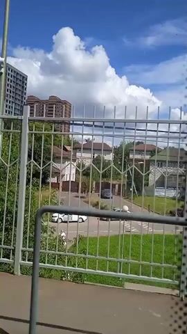 The video opens with a view of a dark structure with a curved roof. A metal fence runs along the right side of the frame. Behind the fence, there are trees and some buildings. The sky is blue with white clouds. The camera then pans to the right, revealing a white metal fence with a decorative pattern. Through the fence, lush green trees are visible, along with some houses and a multi-story building under construction in the distance. A person wearing a blue headscarf and a backpack walks by. Further down the fence, a person wearing a blue t-shirt and a cap walks by, carrying a colorful shopping bag. The camera continues to pan, showing more of the fence, trees, and buildings. The overall environment appears to be an urban or suburban area on a sunny day.