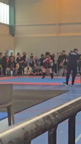 The video shows two young individuals engaged in a martial arts match on a blue and red mat in a large indoor gymnasium. Both fighters are wearing protective gear including helmets, gloves, and shin guards. One fighter wears a pink top, while the other is in a black and yellow outfit. A crowd of spectators surrounds the mat, with a referee overseeing the match. 

In the foreground, a metal barrier runs along the bottom of the frame, partially obscuring the view. A chair sits nearby. A person wearing a black shirt with the words "COACH BEUVRAGES" on the back approaches one of the fighters with a green container in hand. The environment appears to be well-lit with a high ceiling and a basketball hoop visible in the background.
