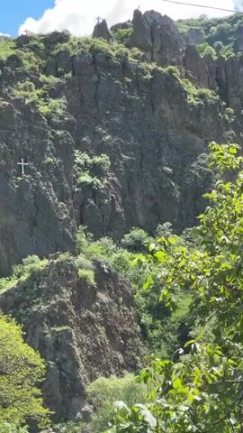 The video opens with a static shot of a steep, rocky cliff face covered in sparse green vegetation. A large white cross is affixed to the left side of the cliff, and several smaller white crosses are visible on the jagged peaks at the top. A thin wire or cable stretches horizontally across the upper portion of the frame against a blue sky with some clouds. The camera then pans down to reveal a crowded outdoor scene. In the immediate foreground, the backs of the heads and shoulders of several individuals are visible, including one person wearing a white sun hat and another with a dark blue head covering, appearing to observe the activity below. Further down, a mix of market stalls with colorful awnings (red, blue) displaying goods is on the left, while a parking lot filled with numerous parked cars (silver, red, dark green, dark blue, white) occupies the right side. People are walking and congregating throughout the market and parking area. The entire scene is surrounded by abundant green trees. A dark-colored flag is partially visible on the far right.