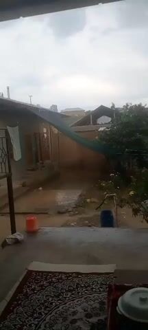 The video shows a backyard during rainfall. Water is visible on the ground and flowing in rivulets. There is a covered walkway or patio area with laundry hanging on a line. A plastic water pump is present, with a blue barrel beside it. In the background, houses and trees are visible under a cloudy sky. The rain appears to be heavy, as evidenced by water cascading from the edge of a roof.