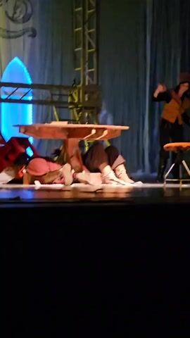 A stage performance is depicted in the video. In the foreground, a person in dark clothing is lying under a wooden table. They are wearing boots and have fabric draped around them. A second person, dressed in a vest and hat, stands to the right of the table, holding a broom. They are seated on a stool. The background features a blue curtain and a metal scaffolding structure.