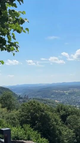 This is a vertical video recording of a panoramic landscape view from a high vantage point on a sunny day. The sky is a clear blue with scattered white clouds. In the foreground, there are lush green trees and foliage, with the tops of some branches and leaves visible on the left side of the frame. Below the trees, a vast valley unfolds, filled with rolling hills, diverse vegetation, and a town or city with buildings. A large body of water, likely a lake or river, is visible in the middle ground of the landscape. In the distance, more hills and a hazy horizon can be seen. A stone wall or barrier is partially visible at the bottom of the frame, suggesting the viewpoint is from a balcony, terrace, or overlook. The video pans slowly to the right, revealing more of the expansive landscape. No people or animals are visible in the recording.