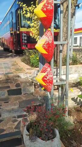 A vertical shot shows a metal pole with a stack of Vietnamese conical hats, also known as nón lá, hanging on it. The hats are red with yellow triangular patterns. In the background, a red and yellow bus is parked, and there are trees and buildings visible. The sky is blue with some clouds. The hats are decorated with small red flowers and yellow flowers, and there is a light fixture attached to the pole.