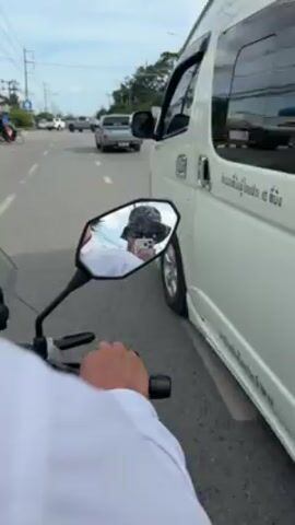 A first-person perspective from a motorcycle traveling on a road, the rider wearing a white shirt and holding the handlebar. In the side mirror, a person wearing a dark patterned hat and sunglasses takes a selfie with a white phone. Cars and trucks are visible on the road. The sky is cloudy with patches of blue. The motorcycle is moving beside a white van.