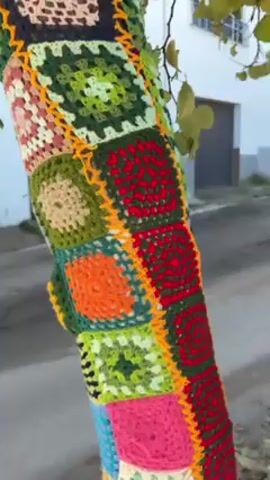 The camera pans upward on the trunk of a tree that is adorned with a multicolored crochet covering made of stitched-together granny squares. The squares are various colors including red, pink, orange, beige, blue, and several shades of green. The leaves of the tree are visible, as is the curb of the road. A white house with a grey garage door sits in the background.