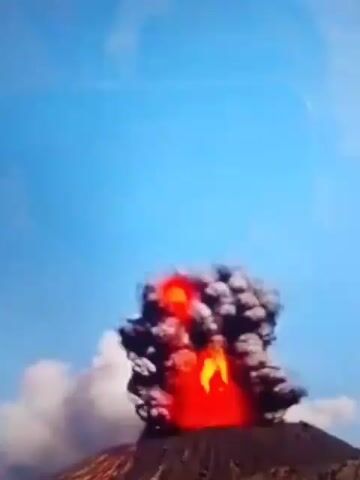 The video captures a volcanic eruption. A large plume of ash and smoke billows into a clear blue sky, with vibrant red and orange lava spewing from the volcano's peak. The eruption appears to be forceful, with molten rock and ash being ejected high into the atmosphere. The surrounding landscape is dark and rocky, indicative of volcanic terrain. The video is taken from a distance, allowing a full view of the dramatic geological event.
