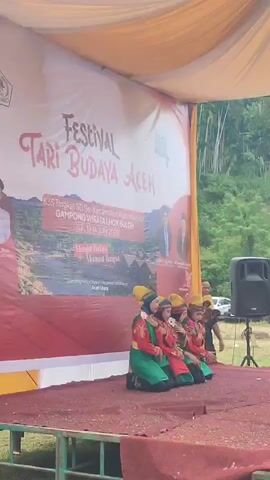A banner reading "Festival Tari Budaya Aceh" is displayed on a stage. Five young performers in traditional Acehnese attire are kneeling on the stage. The performers are wearing colorful costumes with yellow headwear, red tops, and green bottoms. The stage is covered with a red carpet and is set up outdoors under a yellow canopy. In the background, there are trees and a speaker system. A man is visible standing behind the performers.