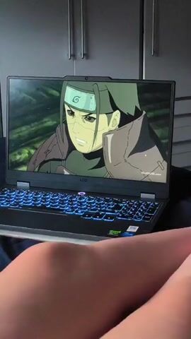 The video shows a laptop screen displaying an anime scene with a male character with dark hair and a headband. The scene abruptly changes to a close-up of a purple flower with green leaves. The laptop is positioned on a surface that appears to be a bed, with a person's legs visible in the foreground. The background shows a closet with two doors. The laptop has a glowing blue keyboard.
