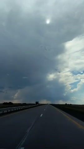 The video shows a view from a vehicle traveling on an empty highway under a dramatic sky. Dark, heavy clouds dominate the upper portion of the frame, with a hint of sunlight peeking through near the top center. Lighter, more diffused clouds are visible towards the right side of the sky, suggesting a storm is approaching or has just passed. The highway stretches out ahead, empty except for a single truck visible in the distance on the left lane. The road has clear white lane markings and yellow edge lines. The surrounding environment is flat and appears to be a rural or open landscape, with a guardrail visible along the left side of the road. The overall impression is one of vastness and movement.
