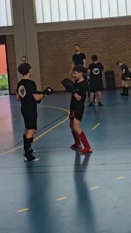The video shows two young people practicing what appears to be kickboxing in a gymnasium. They are wearing black shirts with a logo, black shorts, boxing gloves, and shin guards (one in red). They are facing each other, exchanging practice punches in a sparring-like setup. A coach or instructor is standing in the background, observing the activity. There are other individuals in the background, also dressed in similar attire. The gymnasium has a blue floor with yellow lines, a brick wall, and a window letting in natural light. There is sports equipment along the wall.