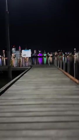 The video shows a person standing on a wooden pier at night, with a city skyline illuminated in the background. The pier is made of wooden planks and extends into the water. There are wooden pilings along the sides of the pier. In the distance, a city skyline is visible with buildings lit up in various colors. The lights reflect on the water, creating a shimmering effect. The person holds a lit cigarette in their right hand and a pink lighter in their left hand. They then move the cigarette out of the frame and walk down the pier towards the city.