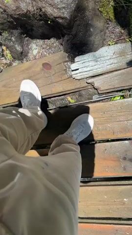 The video begins with a low-angle view of a person's legs and feet, clad in white sneakers and tan pants, standing on a weathered wooden boardwalk. Water trickles down from moss-covered rocks above, onto the boardwalk. The camera then pans up to reveal a cascading waterfall. Large, dark, wet rocks are visible on either side of the waterfall. A dark metal pole is present in the foreground, partially obscuring the view of the falling water. The waterfall crashes into a pool of white, frothy water at its base. The camera continues to ascend, showing more of the powerful waterfall, with greenery and trees visible at the top of the frame.
