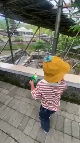 A young child wearing a striped shirt, jeans, white shoes, and a yellow hat adorned with flowers walks along a paved pathway beneath a metal-framed covered walkway. The child is carrying a small, green object in their hand. The walkway overlooks a series of artificial ponds or reservoirs with small waterfalls.  In the background, structures resembling traditional buildings or a temple are visible, along with other trees and greenery. Other people can be seen walking on the pathway. The child points towards the scenery.